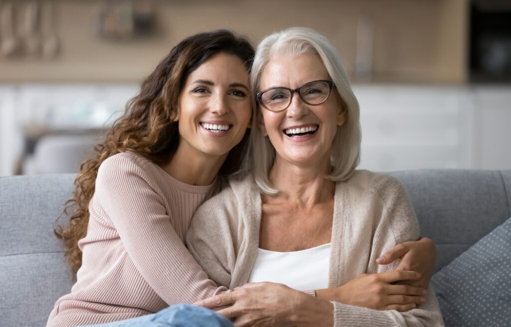 Dental Implants Guide: What to Know Before Replacing Teeth 10 Portrait of two beautiful young and mature females, different generations women hugging sit on sofa, sharing warmth, joyful moments together. Family ties, intergenerational connection, love, support